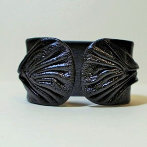Puede incluir: Un brazalete de cuero negro con un dise&ntilde;o &uacute;nico y texturado. El brazalete tiene una banda ancha y plana con dos piezas grandes, redondeadas y texturadas a cada lado.