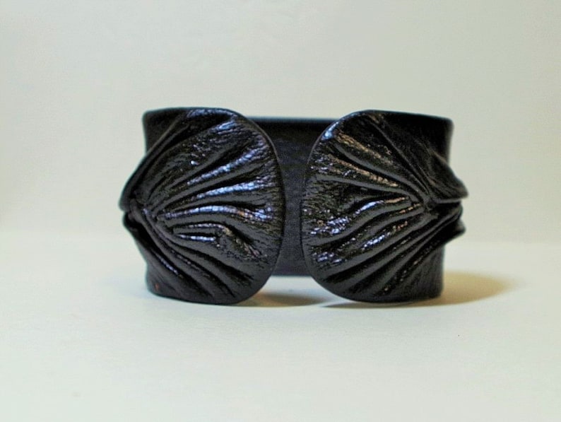 Puede incluir: Un brazalete de cuero negro con un dise&ntilde;o &uacute;nico y texturado. El brazalete tiene una banda ancha y plana con dos piezas grandes, redondeadas y texturadas a cada lado.