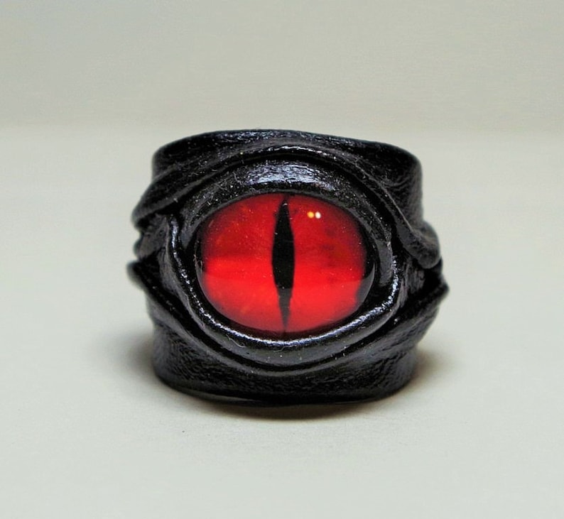 Peut inclure: Une bague en cuir noir avec un motif d'&oelig;il rouge. La bague est en cuir et a un &oelig;il rouge au centre.