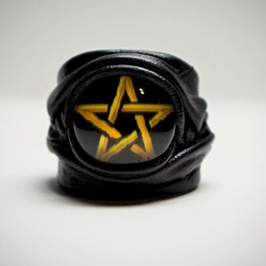 May include: Black leather ring with a gold pentagram design. The ring is made of braided leather and has a round, glass cabochon with the pentagram design.