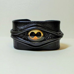 Puede incluir: Pulsera de cuero negro con un dise&ntilde;o de ojo dorado y negro.