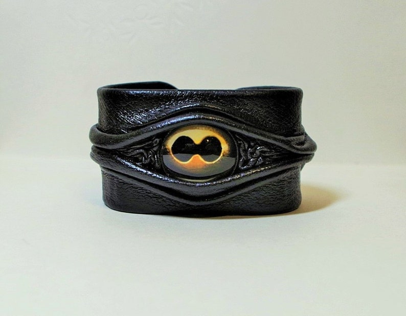 Puede incluir: Pulsera de cuero negro con un dise&ntilde;o de ojo dorado y negro.