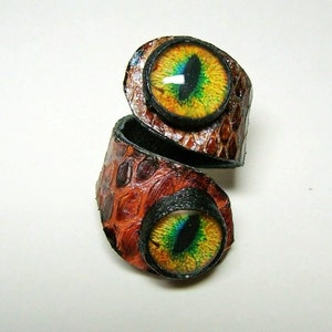 Może przedstawiać: Pierścień skórzany z dwoma dużymi, kolorowymi szklanymi oczami. Pierścień jest brązowy z fakturowanym wzorem i jest owinięty wokół siebie.