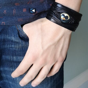 Puede incluir: Pulsera de cuero negro con un dise&ntilde;o de ojo dorado y negro.