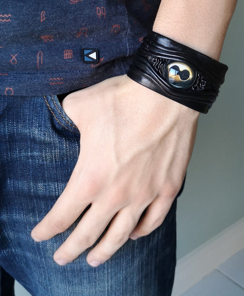 Puede incluir: Pulsera de cuero negro con un dise&ntilde;o de ojo dorado y negro.
