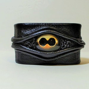 Puede incluir: Pulsera de cuero negro con un dise&ntilde;o de ojo decorativo. El ojo es amarillo y naranja con una pupila negra.