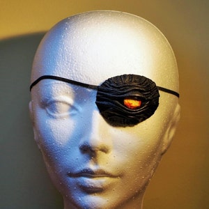 May include: A black leather eye patch with a textured surface and a red and orange eye detail. The eye patch is worn over a white mannequin head.