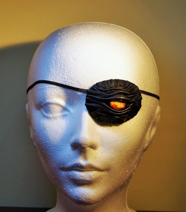 May include: A black leather eye patch with a textured surface and a red and orange eye detail. The eye patch is worn over a white mannequin head.