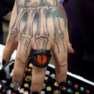 Peut inclure: Une bague en cuir noir avec un motif d'&oelig;il rouge. La bague est port&eacute;e sur le majeur d'une main avec un tatouage de squelette.