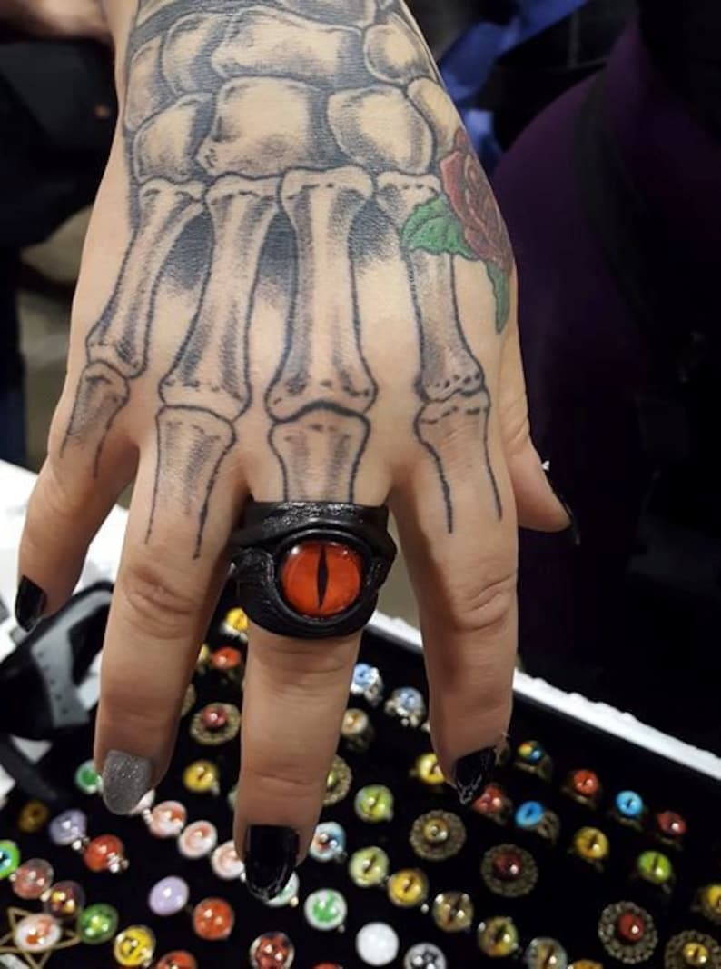 Peut inclure: Une bague en cuir noir avec un motif d'&oelig;il rouge. La bague est port&eacute;e sur le majeur d'une main avec un tatouage de squelette.