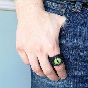 Peut inclure: Une bague en cuir noir avec un motif d'&oelig;il vert. La bague est port&eacute;e sur le majeur d'une main.