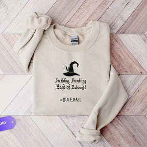 May include: A sand-colored sweatshirt with a crew neck and rolled-up sleeves. The front features a black witch hat graphic and the text "Babbling, Bumbling Band of Baboons!" and "#YULEBALL". The sweatshirt is on a wooden surface.