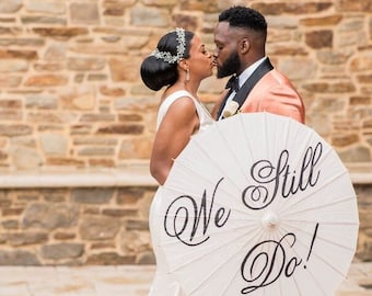 May include: A bride and groom in formal attire are kissing in front of a stone wall. They are holding a white umbrella with the words "We Still Do!" written in black.