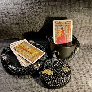 Op de afbeelding: Zwarte slangvormige doos met een deksel en een rond deksel met gouden accenten. De doos is open en bevat een deck tarotkaarten. De bovenste kaart is de 5 van Staven.