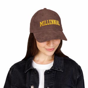 Puede incluir: Gorra de béisbol de pana marrón con la palabra "MILLENNIAL" bordada en amarillo. La gorra tiene una visera curva y un botón en la parte superior. La persona lleva una chaqueta vaquera negra.