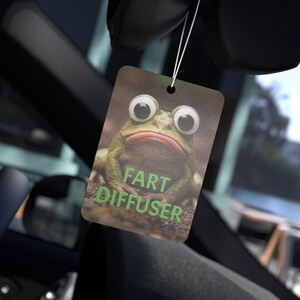 Puede incluir: Un ambientador rectangular para coche con un diseño de rana y las palabras "FART DIFFUSER" en verde. La rana tiene grandes ojos saltones y una boca roja. El ambientador cuelga de una cuerda.