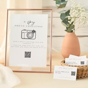 May include: A framed sign reading "I Spy Photo Challenge" with a camera illustration, alongside cards in a wicker basket. A vase of flowers and a white cloth are also in the image. The sign includes a QR code.