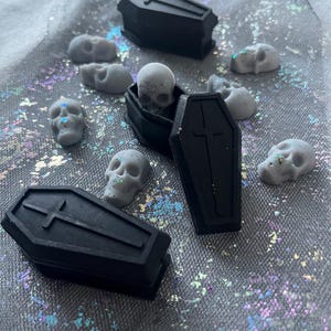 May include: A collection of small black coffin-shaped boxes with a cross on top. Each box contains a small grey skull. The boxes and skulls are on a grey and white sparkly surface.