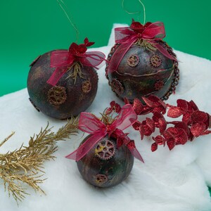 May include: Three dark brown Christmas ornaments with a steampunk design, decorated with gears and red ribbon bows. The ornaments are sitting on a white fluffy surface with a sprig of red glitter leaves.