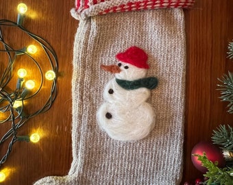 May include: A knitted Christmas stocking with a snowman design. The stocking is beige with a red and white striped cuff. The snowman is white with a red hat, green scarf, and black eyes and buttons.
