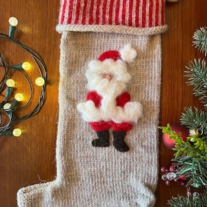 May include: A knitted Christmas stocking with a Santa Claus design. The stocking is beige with a red and white striped cuff. The Santa Claus is made of felt and has a white beard and a red suit.