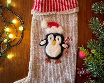 May include: A knitted Christmas stocking with a penguin design. The stocking is beige with red and white stripes at the top. The penguin is made of felt and has a red Santa hat. The stocking has a loop at the top for hanging.