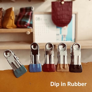 Peut inclure: Cinq pinces à linge en métal argenté avec des embouts en caoutchouc de couleur. Les embouts sont bleus, rouges, dorés et noirs. Le texte "Dip in Rubber" est imprimé sur une surface brune en dessous des pinces.