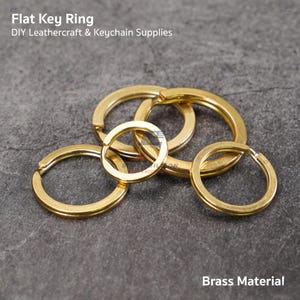 May include: Several gold-colored flat key rings are displayed against a gray background. The key rings are made of brass and are of varying sizes. The text "Flat Key Ring" and "DIY Leathercraft & Keychain Supplies" is visible at the top. The text "Brass Material" is at the bottom.