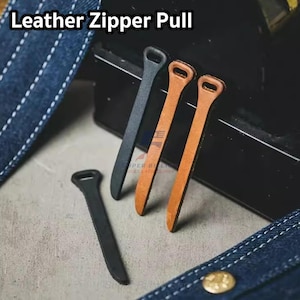 May include: A selection of leather zipper pulls in black and tan are shown. The pulls have a loop at the top and a long, narrow body. The text "Leather Zipper Pull" is at the top of the image. The pulls are displayed on a grey surface.