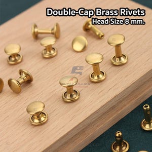 May include: Close-up of brass double-cap rivets with a head size of 8 mm. The rivets are scattered on a wooden surface. The text "Double-Cap Brass Rivets Head Size 8 mm." is visible in the image.