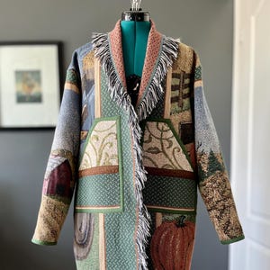 May include: A patchwork tapestry coat featuring rural landscape scenes, including a barn, fence, and pumpkin. The coat has a fringed collar and hem, with green and brown accents. The coat is displayed on a mannequin.