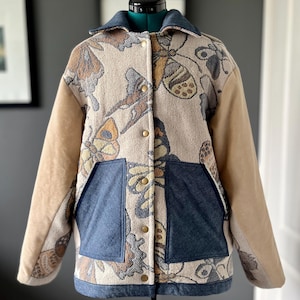 May include: A beige jacket with a brown suede collar and sleeves. The jacket has a button closure and is decorated with a butterfly pattern. The jacket has two large pockets on the front.