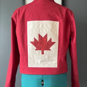 May include: Red jacket featuring a white and red maple leaf design on the back. The maple leaf is made of red glitter. The jacket has long sleeves and a collar. The jacket is displayed on a turquoise dress form.
