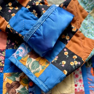 A colourful quilted jacket is displayed on a teal mannequin.  It features a patchwork design with various floral & patterned squares in shades of blue, pink, yellow, orange, and green.  Blue trim outlines the pockets and edges.