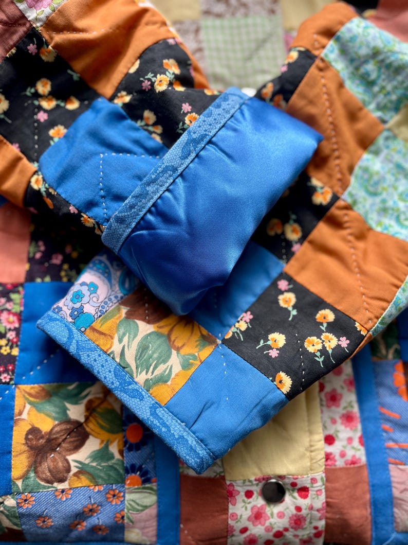 A colourful quilted jacket is displayed on a teal mannequin.  It features a patchwork design with various floral & patterned squares in shades of blue, pink, yellow, orange, and green.  Blue trim outlines the pockets and edges.
