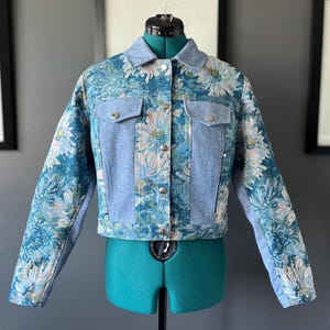 May include: A cropped jacket featuring a mix of light blue denim panels and a floral tapestry fabric in shades of teal, blue, and cream. The jacket includes a classic collar, long sleeves, two chest pockets with flaps, and silver-toned metal buttons.