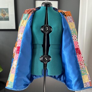 A colourful quilted jacket is displayed on a teal mannequin.  It features a patchwork design with various floral & patterned squares in shades of blue, pink, yellow, orange, and green.  Blue trim outlines the pockets and edges.