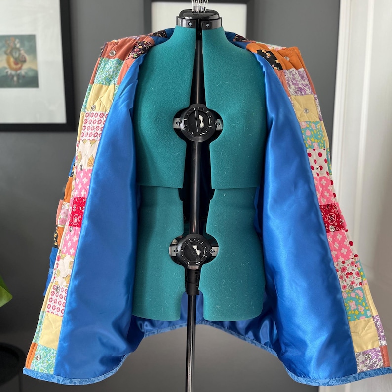 A colourful quilted jacket is displayed on a teal mannequin.  It features a patchwork design with various floral & patterned squares in shades of blue, pink, yellow, orange, and green.  Blue trim outlines the pockets and edges.