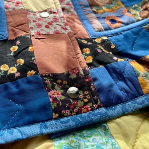 A colourful quilted jacket is displayed on a teal mannequin.  It features a patchwork design with various floral & patterned squares in shades of blue, pink, yellow, orange, and green.  Blue trim outlines the pockets and edges.
