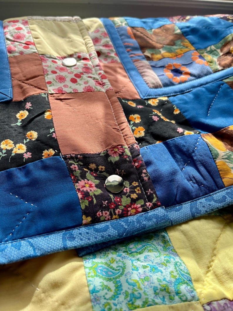 A colourful quilted jacket is displayed on a teal mannequin.  It features a patchwork design with various floral & patterned squares in shades of blue, pink, yellow, orange, and green.  Blue trim outlines the pockets and edges.