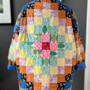 A colourful quilted jacket is displayed on a teal mannequin.  It features a patchwork design with various floral & patterned squares in shades of blue, pink, yellow, orange, and green.  Blue trim outlines the pockets and edges.