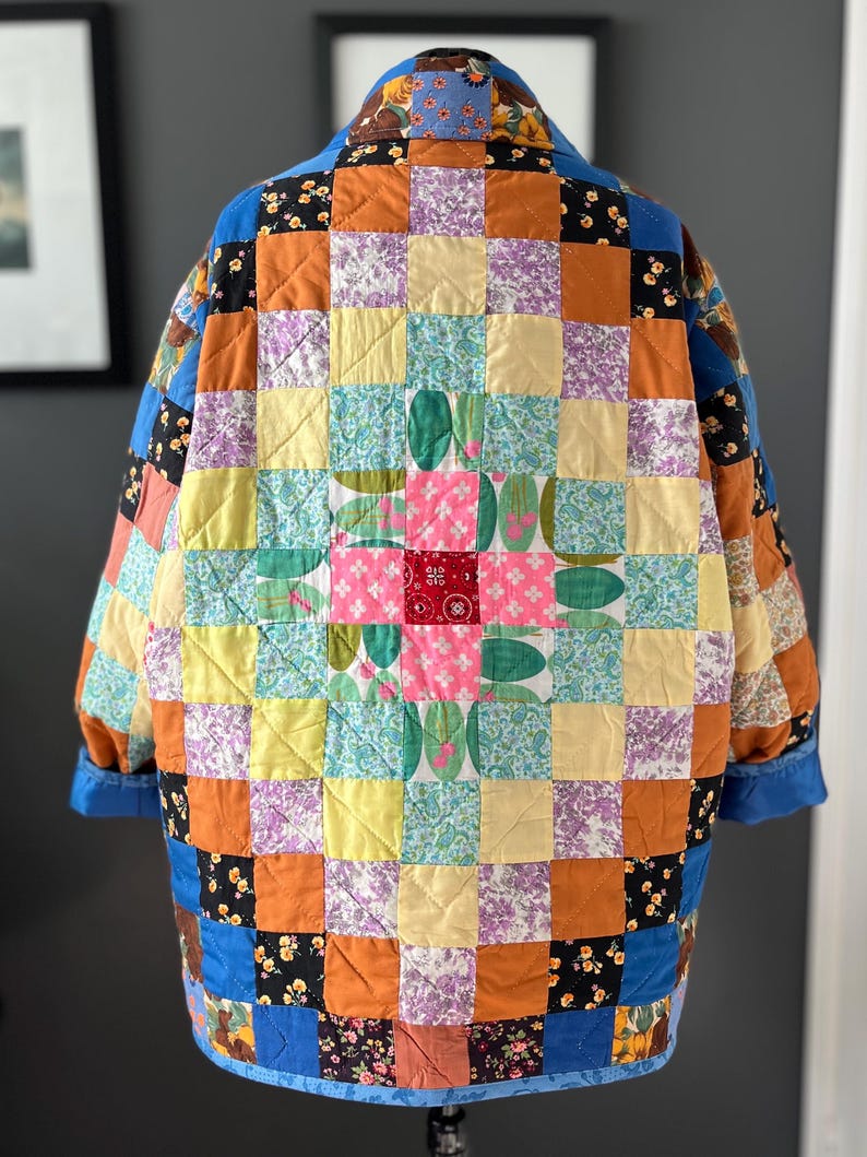 A colourful quilted jacket is displayed on a teal mannequin.  It features a patchwork design with various floral & patterned squares in shades of blue, pink, yellow, orange, and green.  Blue trim outlines the pockets and edges.