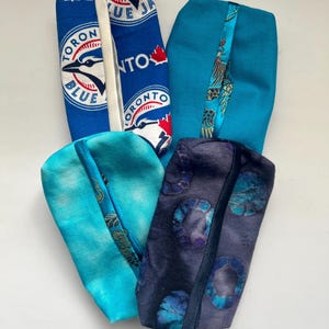 Może przedstawiać: Cztery materiałowe etui w odcieniach niebieskiego. Jedno etui ma logo Toronto Blue Jays w kolorze niebiesko-białym. Pozostałe etui są w jednolitym kolorze niebieskim, z wzorzystymi akcentami. Etui są prostokątne z zakrzywioną górą.