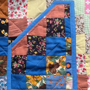 A colourful quilted jacket is displayed on a teal mannequin.  It features a patchwork design with various floral & patterned squares in shades of blue, pink, yellow, orange, and green.  Blue trim outlines the pockets and edges.