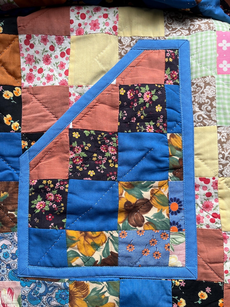 A colourful quilted jacket is displayed on a teal mannequin.  It features a patchwork design with various floral & patterned squares in shades of blue, pink, yellow, orange, and green.  Blue trim outlines the pockets and edges.