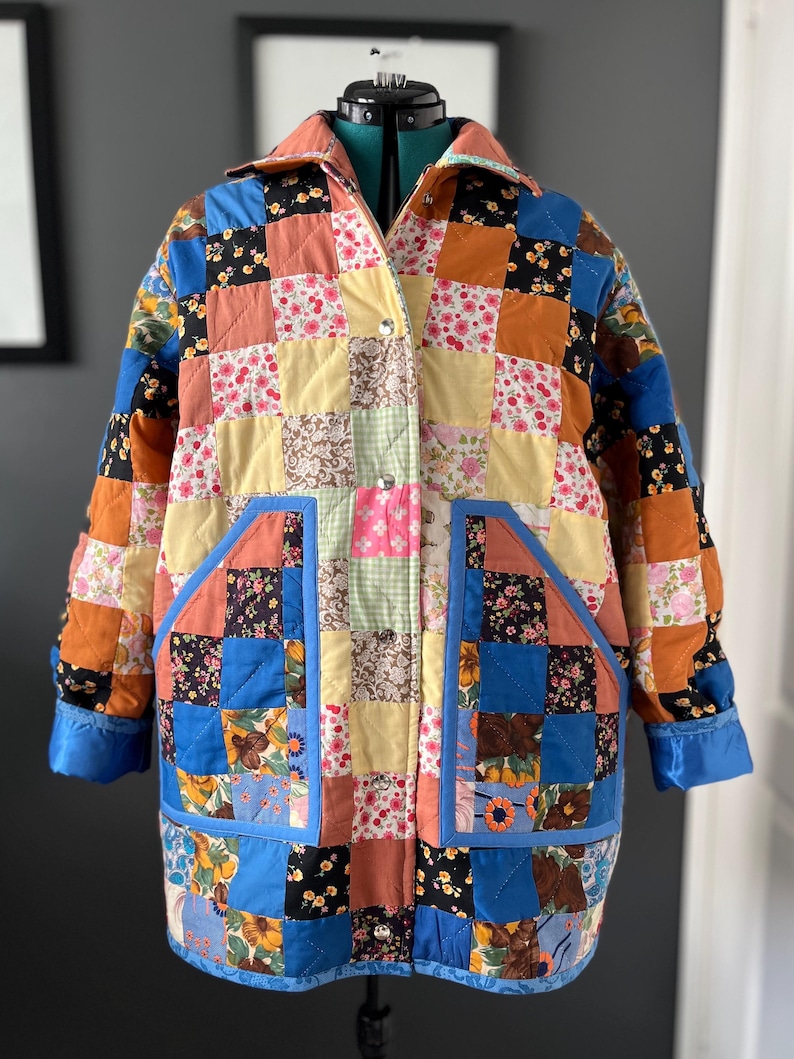 A colourful quilted jacket is displayed on a teal mannequin.  It features a patchwork design with various floral & patterned squares in shades of blue, pink, yellow, orange, and green.  Blue trim outlines the pockets and edges.