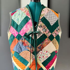 May include: A patchwork quilted vest with a variety of colourful fabric patterns, including floral, polka dot, and geometric designs, set against a dark green background. A green ribbon ties the vest closed.