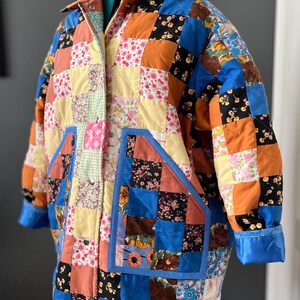 A colourful quilted jacket is displayed on a teal mannequin.  It features a patchwork design with various floral & patterned squares in shades of blue, pink, yellow, orange, and green.  Blue trim outlines the pockets and edges.