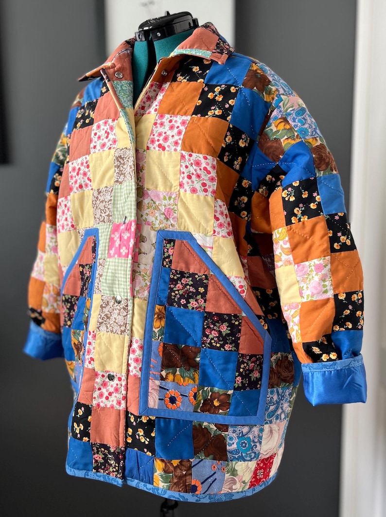 A colourful quilted jacket is displayed on a teal mannequin.  It features a patchwork design with various floral & patterned squares in shades of blue, pink, yellow, orange, and green.  Blue trim outlines the pockets and edges.