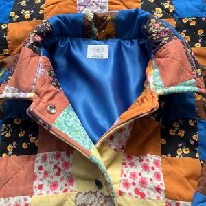 A colourful quilted jacket is displayed on a teal mannequin.  It features a patchwork design with various floral & patterned squares in shades of blue, pink, yellow, orange, and green.  Blue trim outlines the pockets and edges.